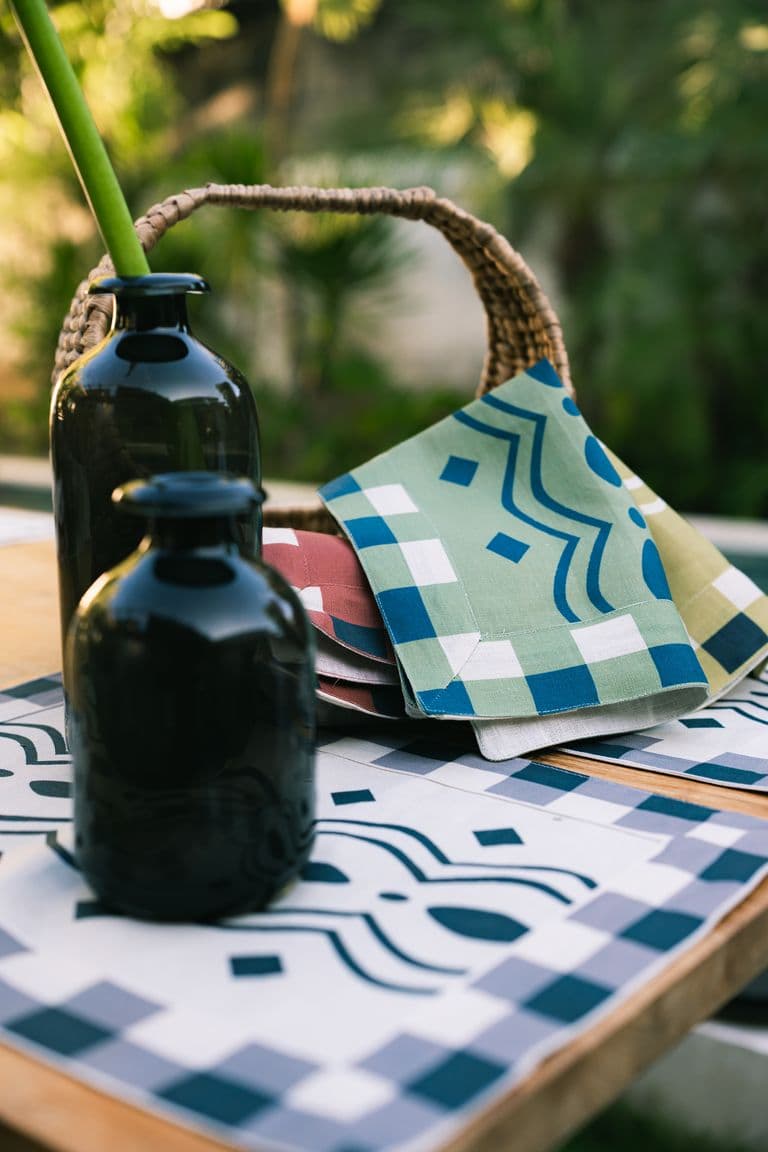 KIND napkins and placemats in green, blue and terracotta prints beside dark glass vases in a garden